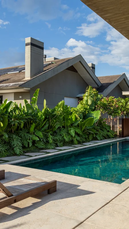 Swimming pool with stone deck next to modern house and lush green plants under blue sky.