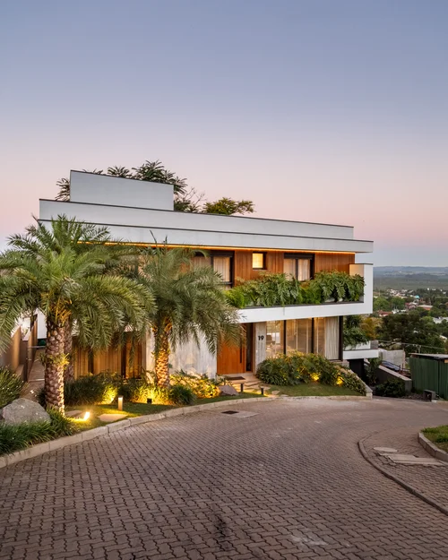Modern two-story house with palm trees and greenery at dusk on a paved curved driveway.