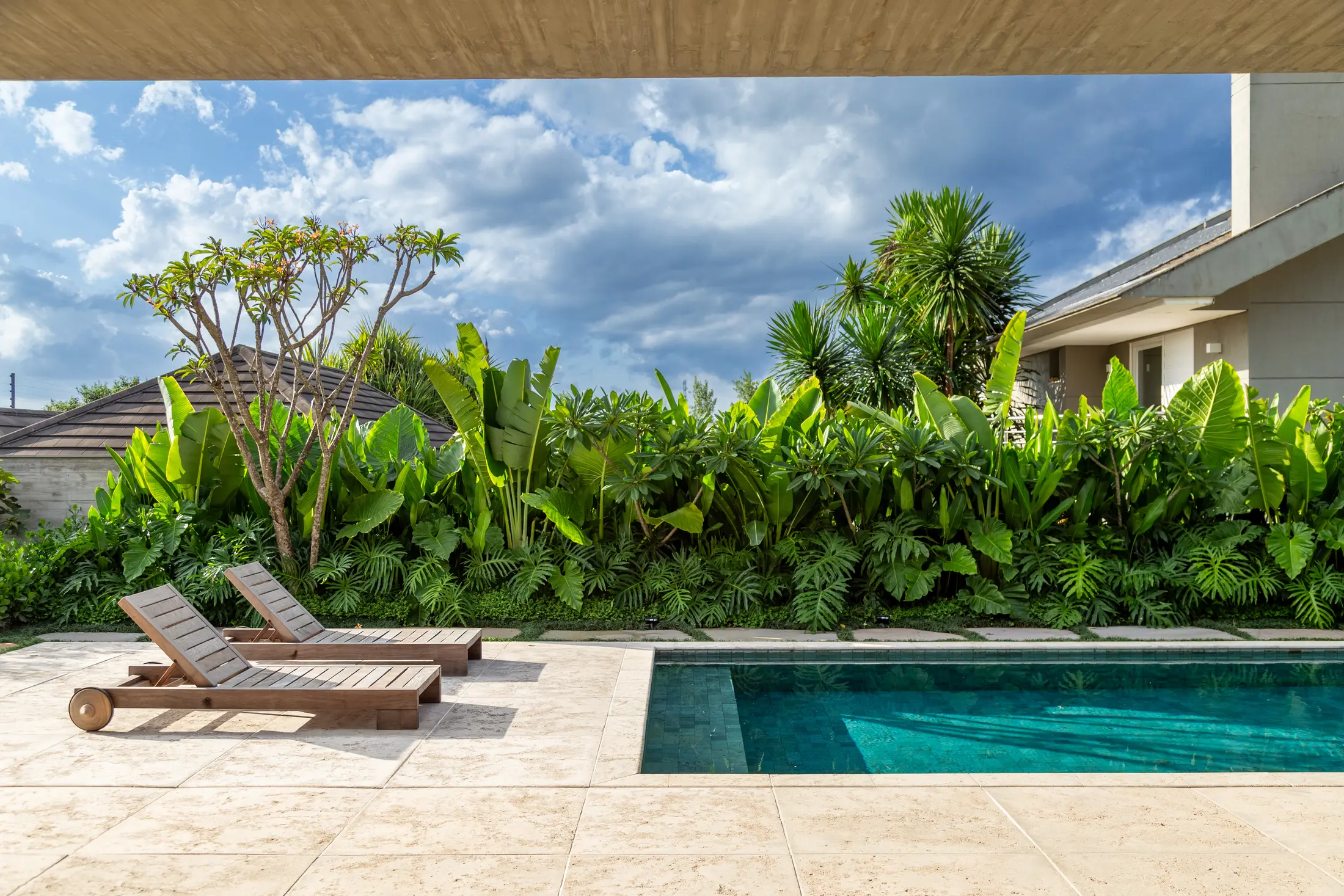 Two wooden lounge chairs by a blue pool with lush tropical plants and a partly cloudy sky.