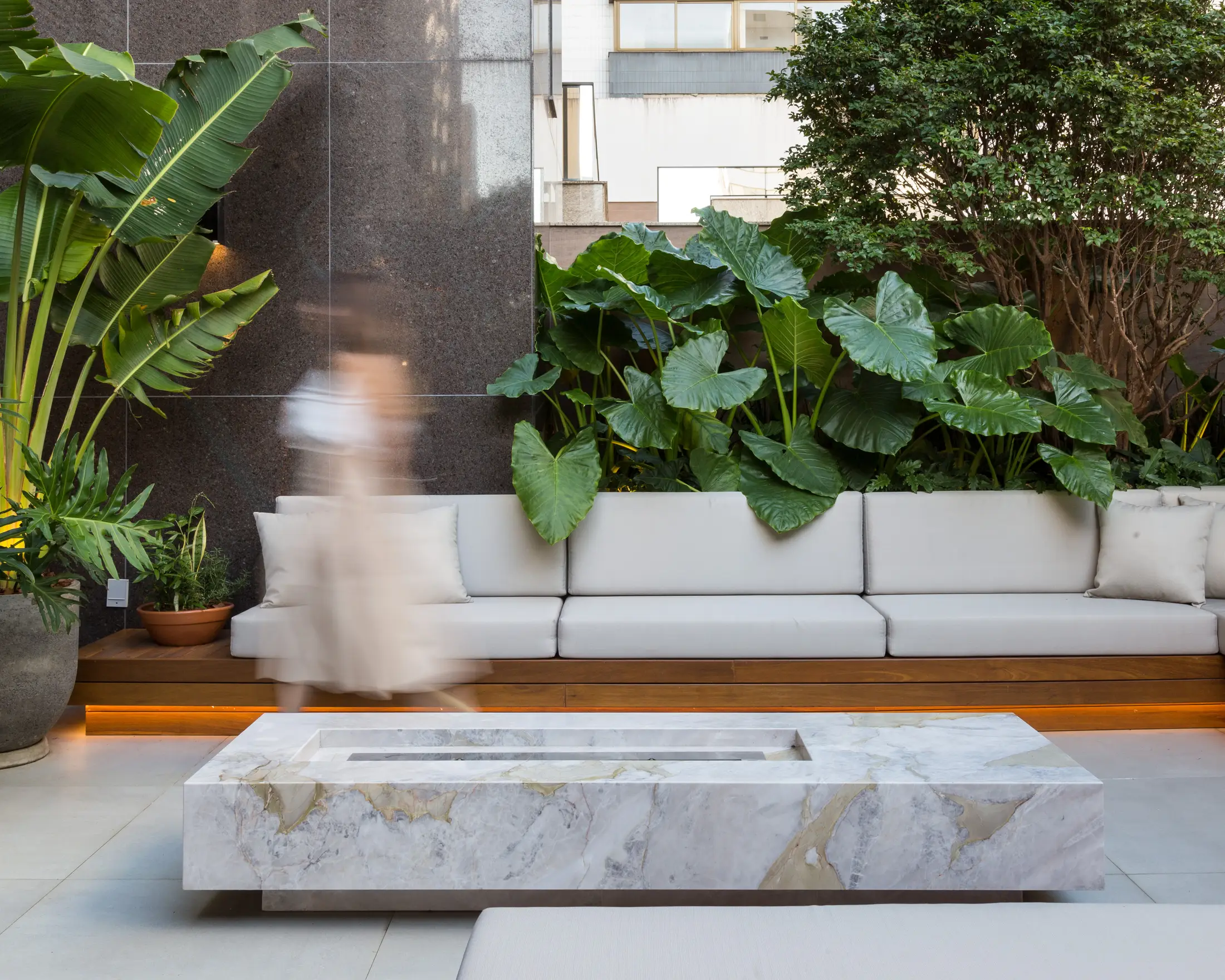 Blurred person walking past a modern outdoor seating area with large green plants and a marble table.