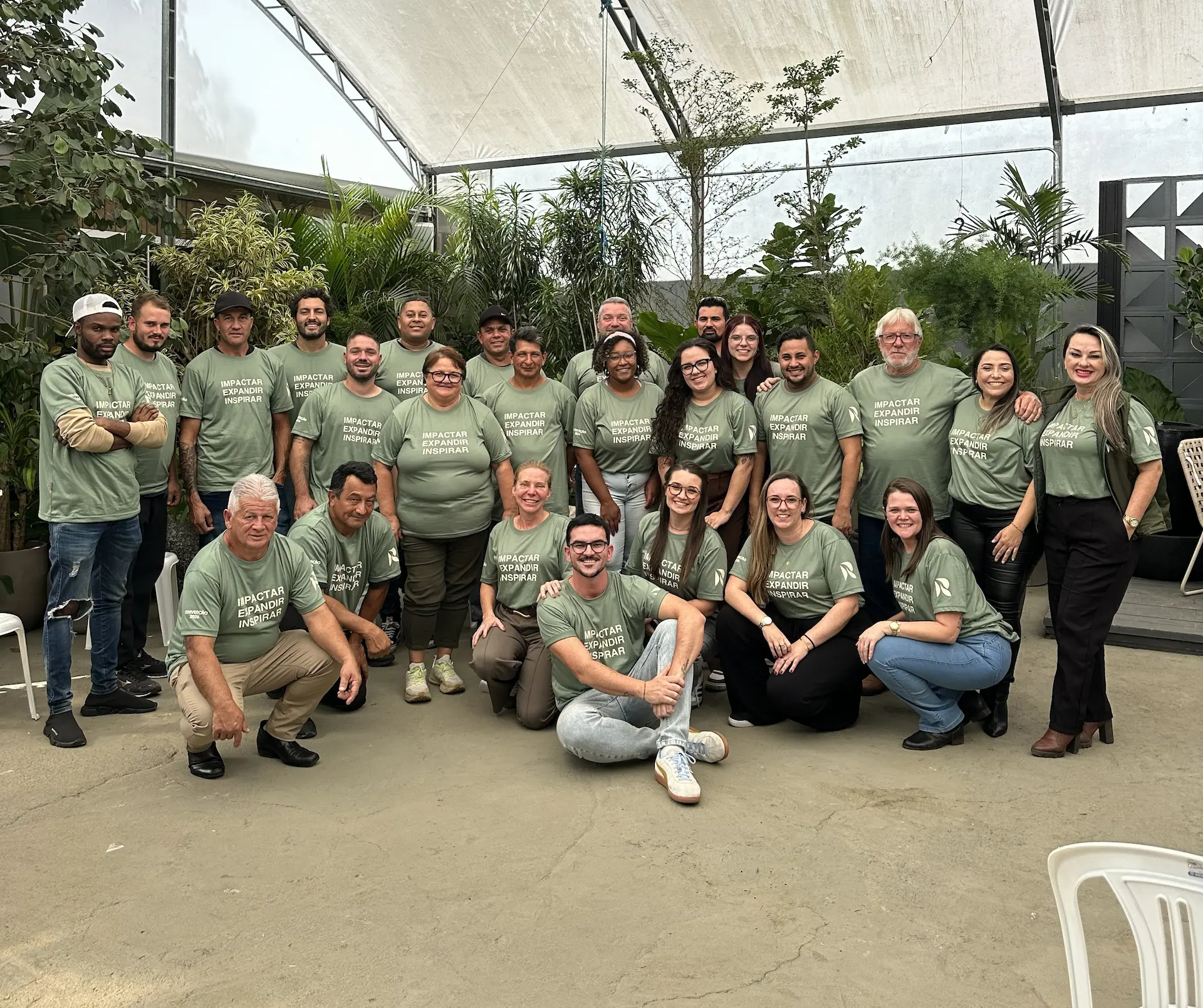 Group of 24 people wearing matching green shirts posing inside a greenhouse with plants.