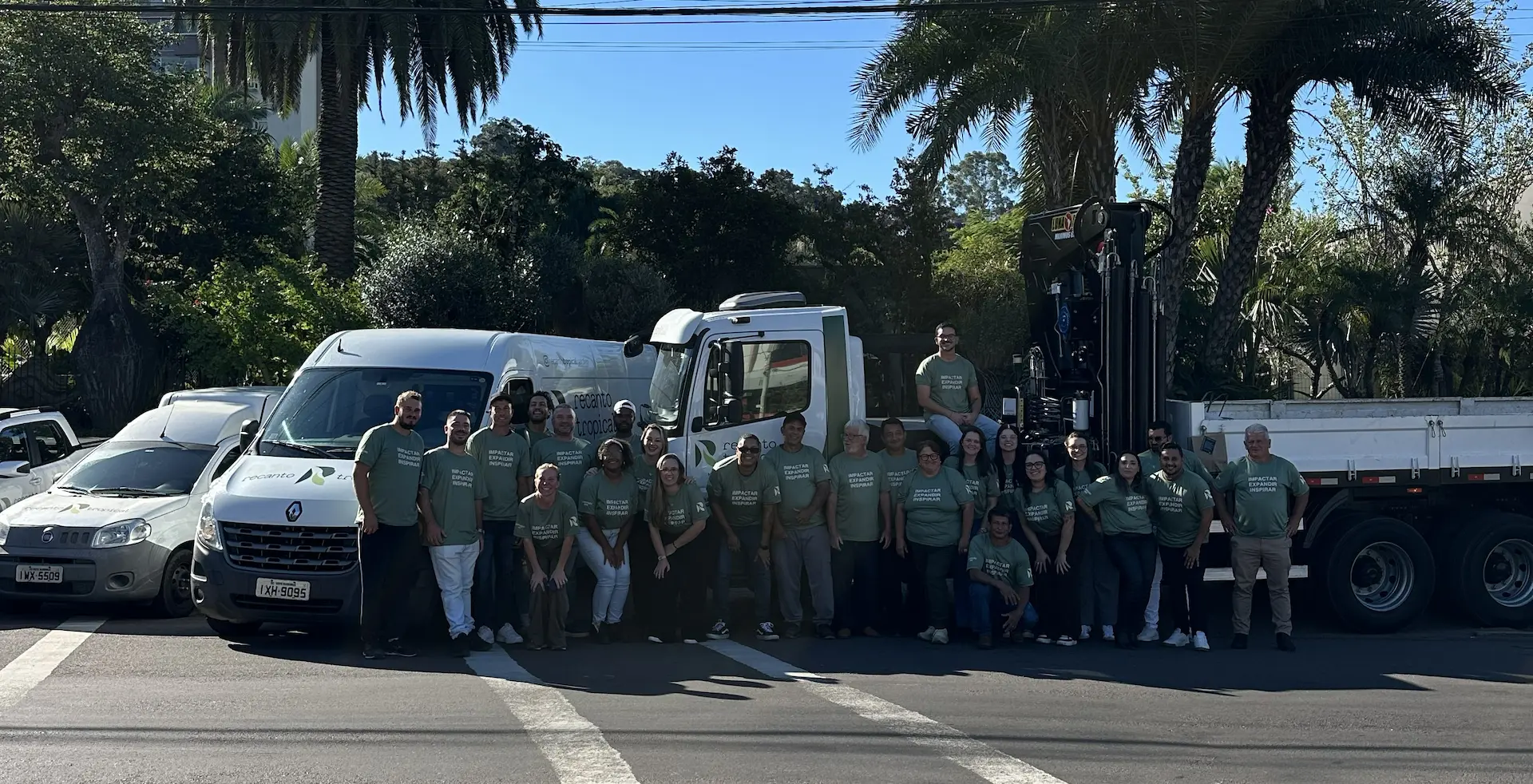 Group of people in matching shirts posing outdoors in front of trucks and vans with tropical plants behind them.