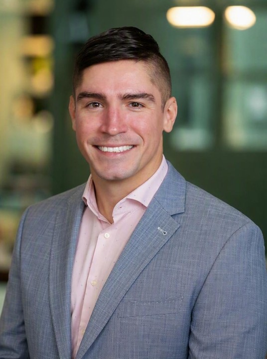 Smiling man with dark hair and styled mustache wearing a light gray suit jacket and pink shirt.