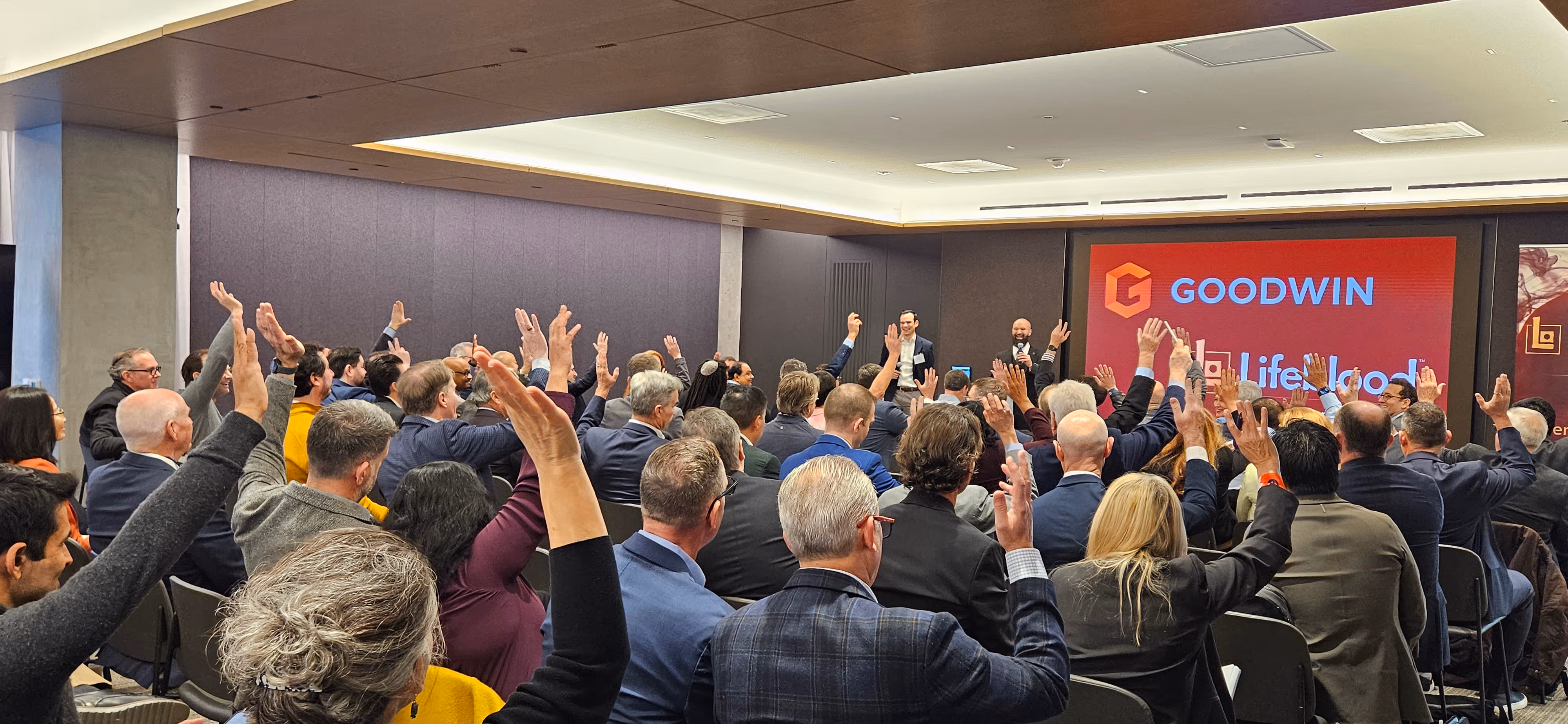 Audience at a business event raising hands while two presenters speak near a screen with Goodwin logo.
