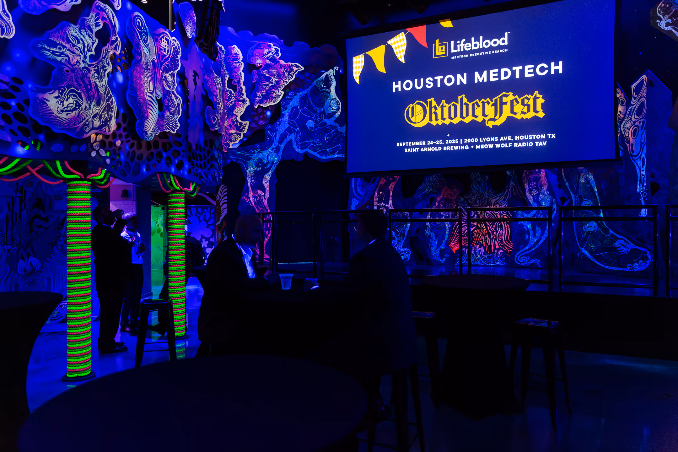 Dimly lit event space with neon decorations and a screen announcing Houston MedTech Oktoberfest.