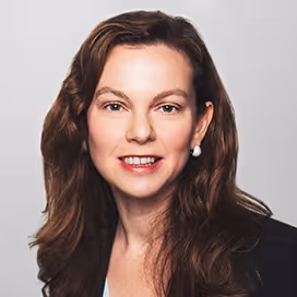 Portrait of a woman with long brown hair wearing pearl earrings and a dark blazer.