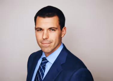 Professional headshot of a man in a navy suit and blue tie against a plain light background.