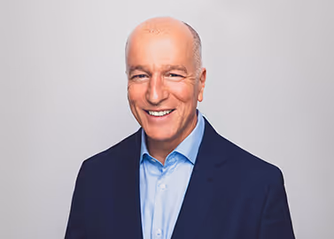 Smiling bald man wearing a blue suit jacket and light blue shirt against a gray background.