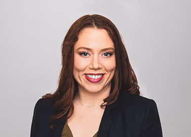 Smiling woman with long brown hair wearing a black blazer and green top against a gray background.