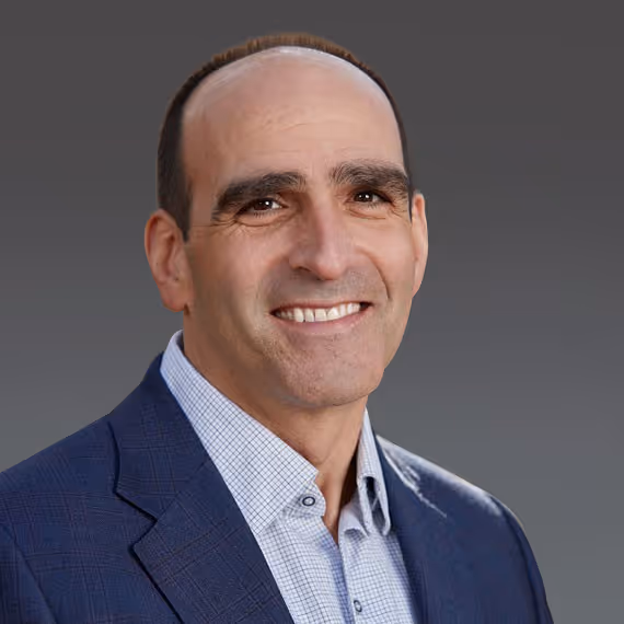 Smiling middle-aged man in a blue suit jacket and checkered shirt against a gray background.