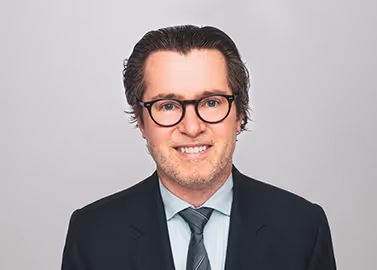 Man with glasses and dark suit smiling against a plain light background.