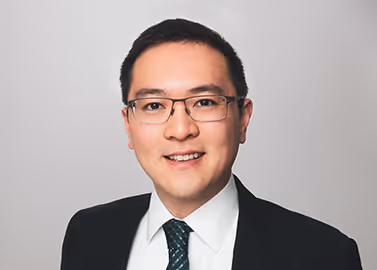 Smiling man in glasses wearing a dark suit, white shirt, and patterned tie on a gray background.
