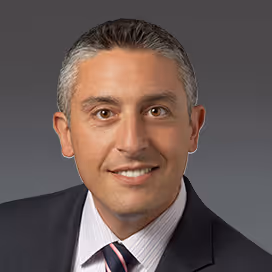 Smiling man with graying hair wearing a dark suit, white shirt, and striped tie on gray background.