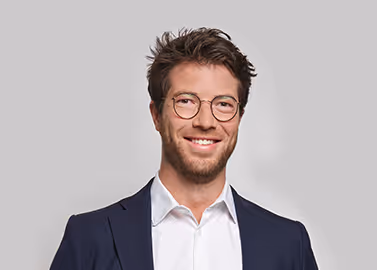 Smiling man with glasses in a white shirt and dark blazer against a gray background.