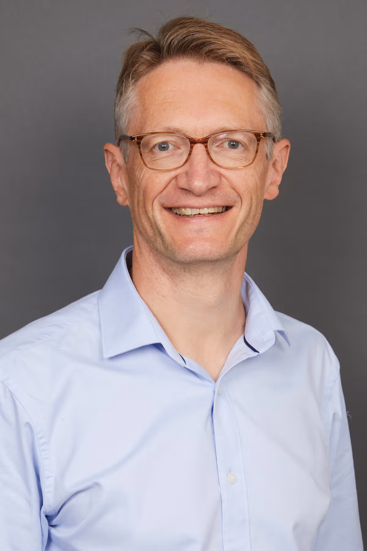 Smiling middle-aged man with light brown hair and glasses wearing a light blue shirt against gray background.