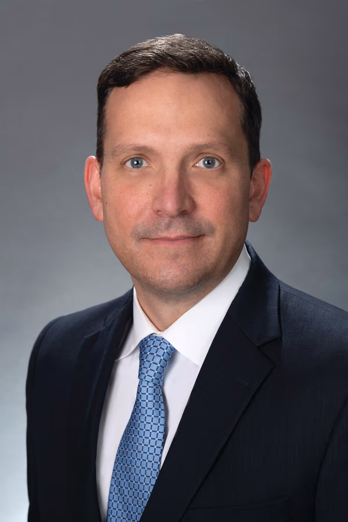 Portrait of a man in a dark suit, white shirt, and blue patterned tie on a gray backdrop.