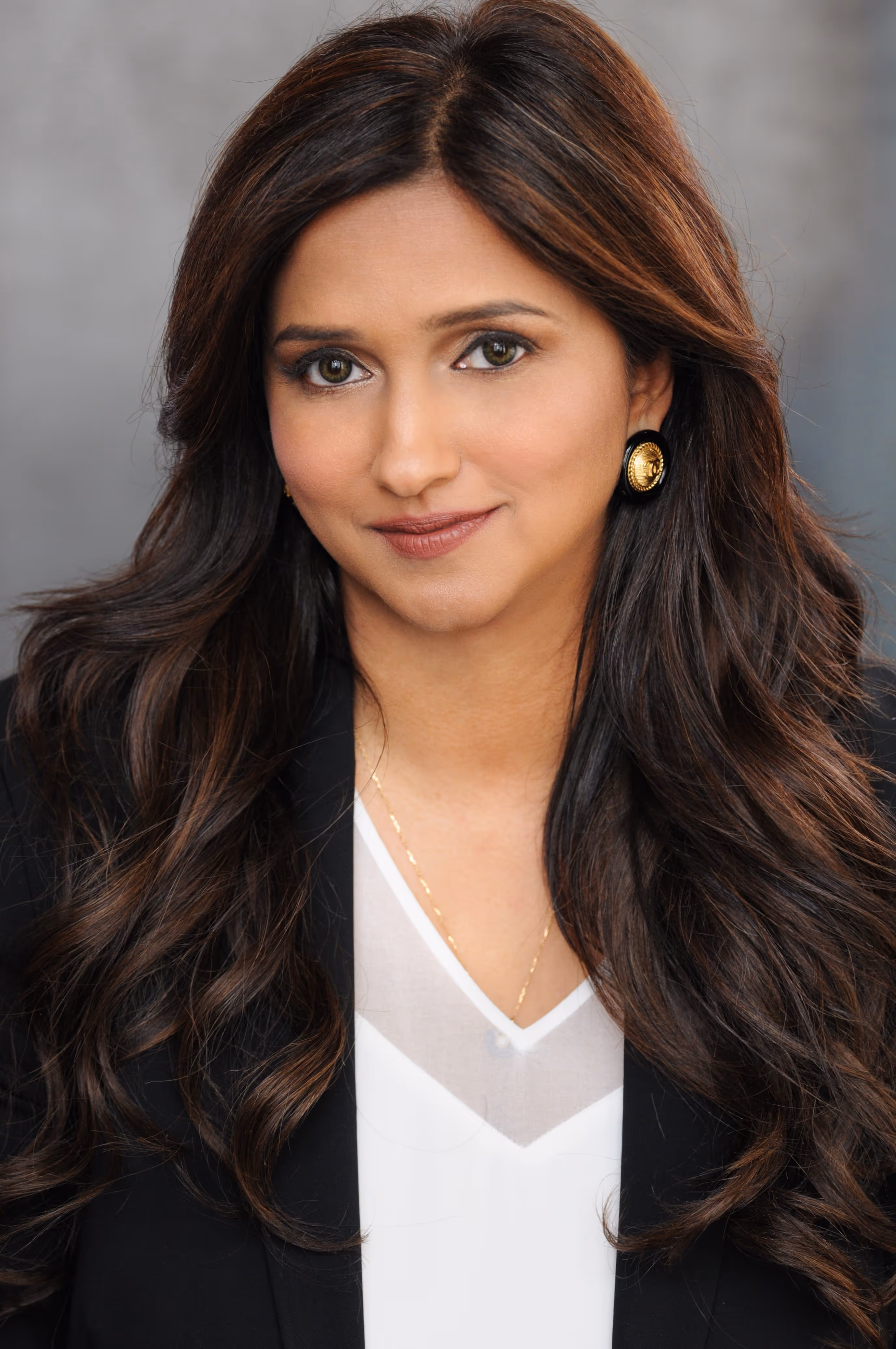 Smiling woman with long dark hair wearing a black blazer, white blouse, and gold earrings.