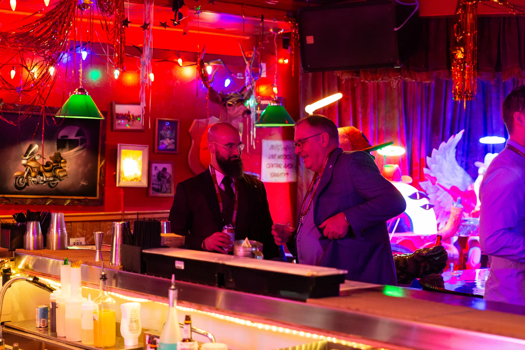 Two men talking and holding drinks at a vibrant bar with colorful lights and eclectic décor.