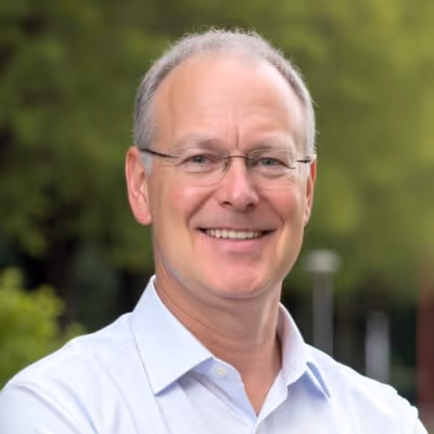 Smiling middle-aged man with glasses wearing a light blue shirt outdoors with blurred green background.