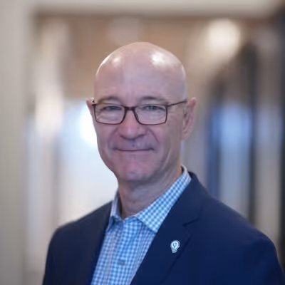 Smiling bald man with glasses wearing a checkered shirt and dark blazer indoors.