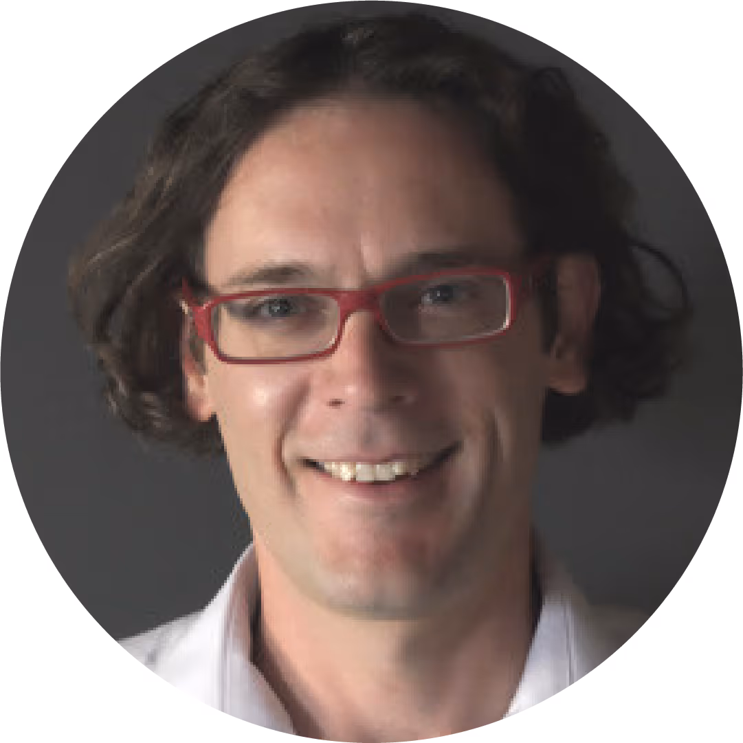 Smiling man with curly brown hair and red glasses wearing a white shirt, gray background.