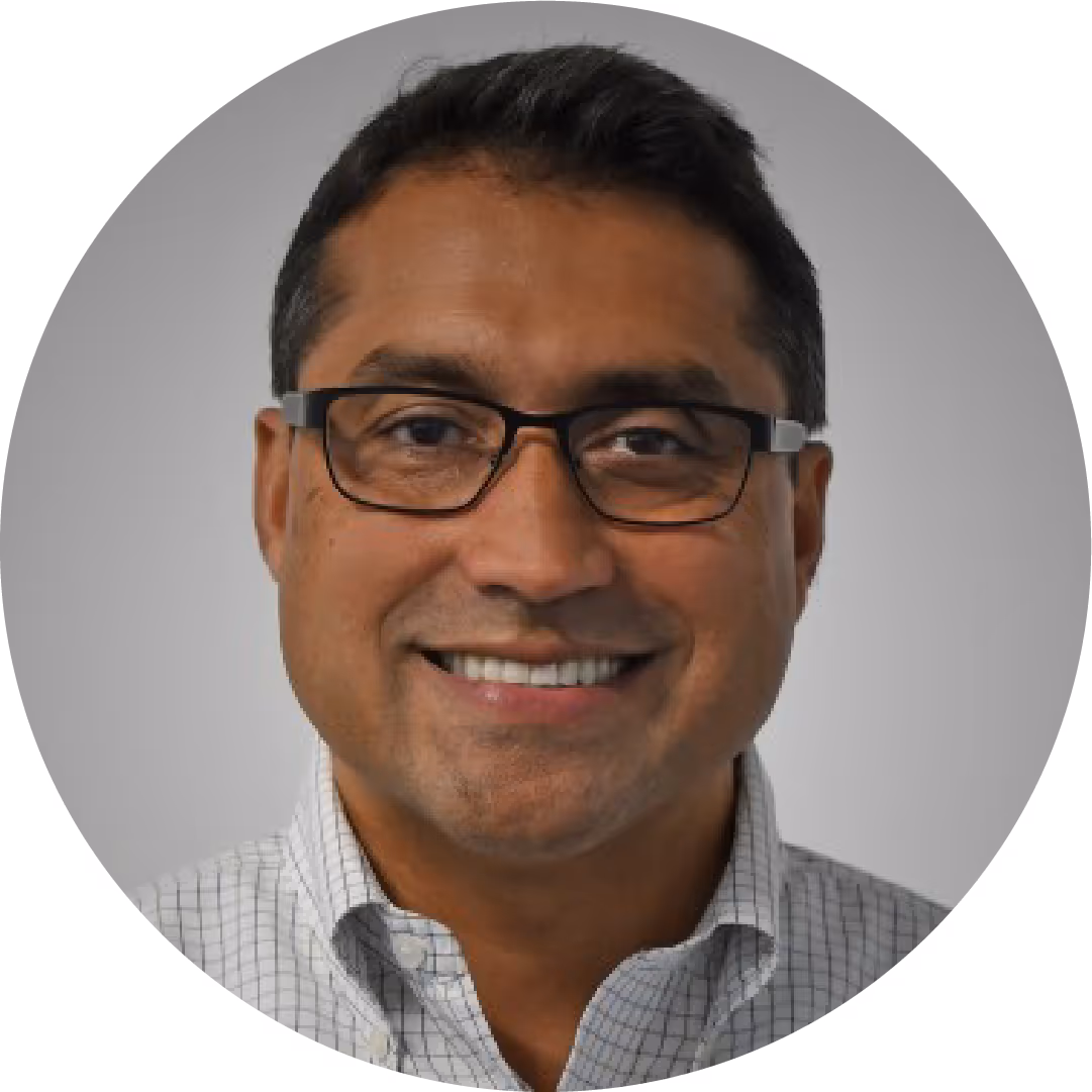 Smiling man with glasses and checkered shirt in front of a gray background.