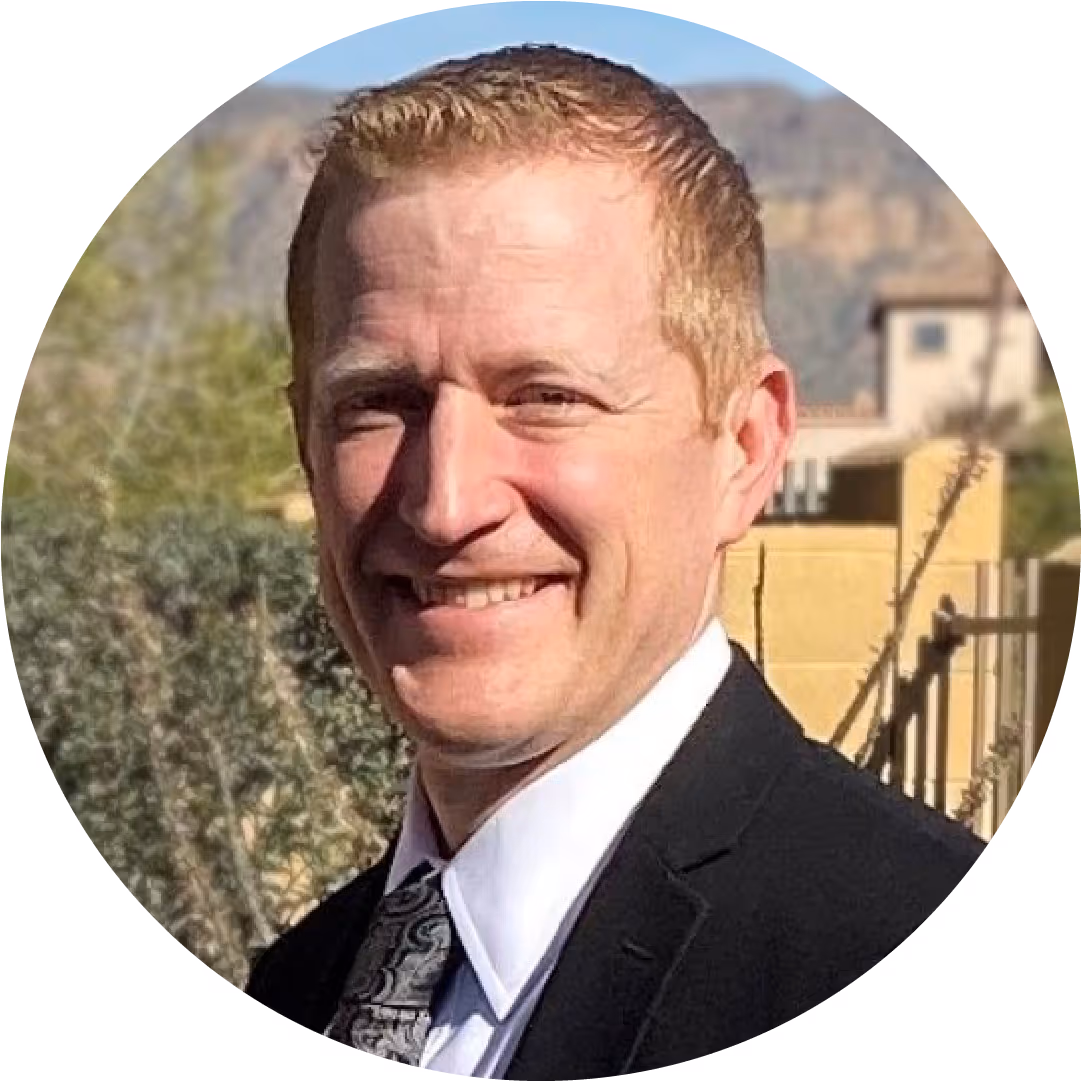Smiling man with short red hair wearing a black suit, white shirt, and patterned tie outdoors.