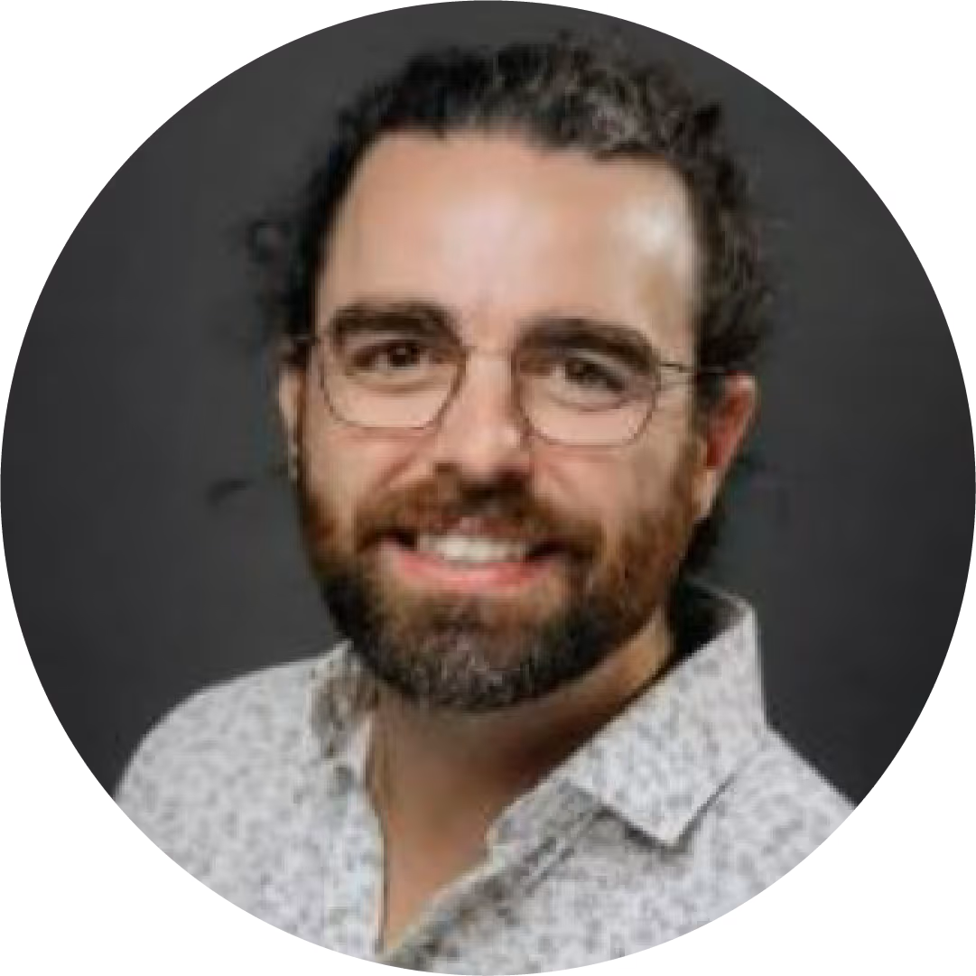 Man with glasses, beard, and curly hair smiling, wearing a light patterned shirt against a dark background.