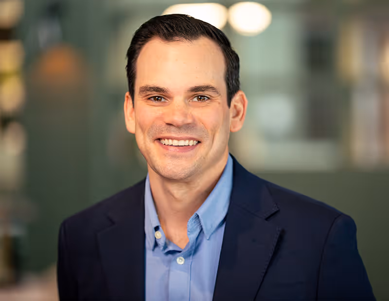 Smiling man with dark hair wearing a blue shirt and dark blazer, blurred indoor background.