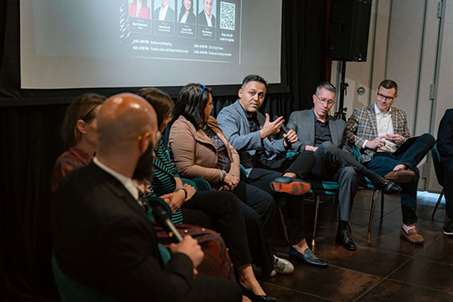 Audience watching panel discussion with four men on stage in rustic venue with string lights.