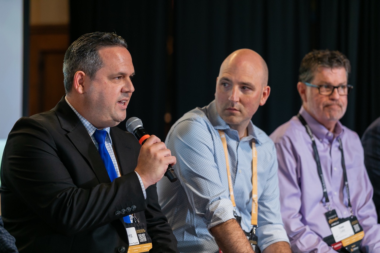 Three men seated indoors, two holding microphones, one speaking during a panel discussion.