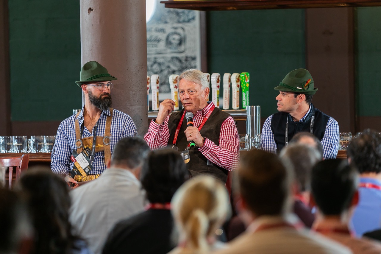 Seven men seated on stools holding microphones and drinks at Houston MedTech Oktoberfest panel event.