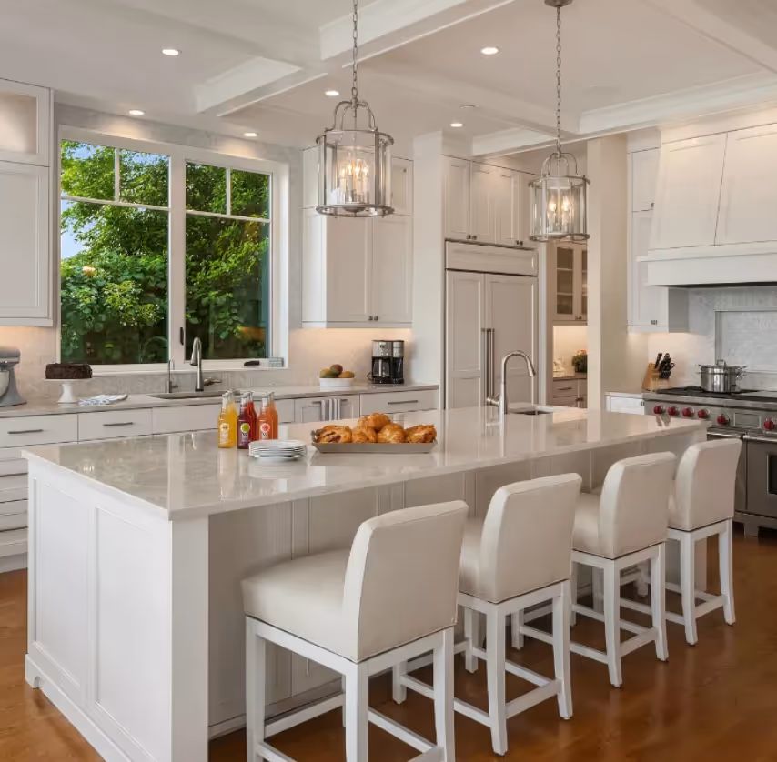 Modern white kitchen island with a bowl of bananas and oranges, gas stove, and a background of plants, stairs, and living area.