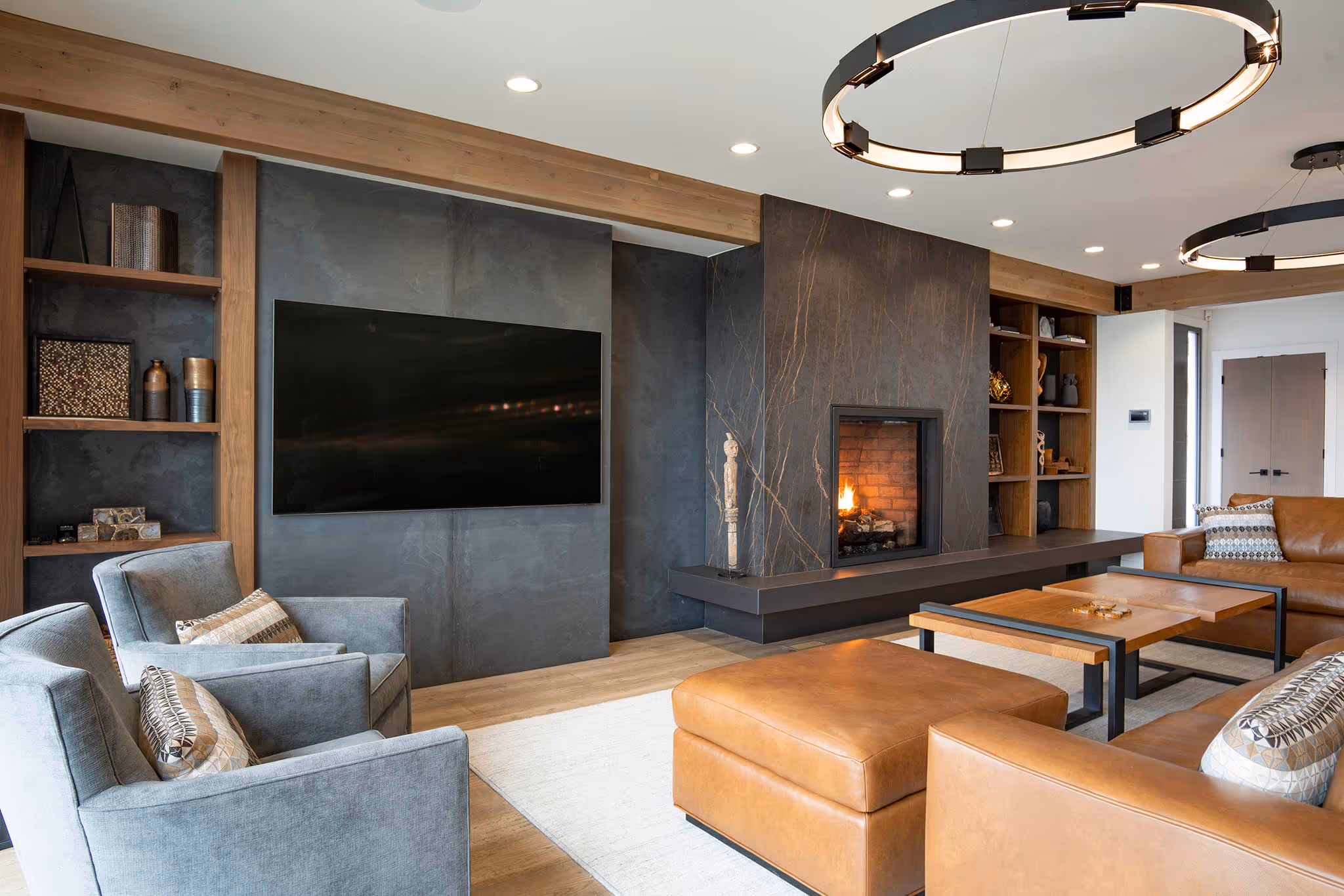 Modern living room with gray armchairs, leather sofas, a wooden coffee table, a wall-mounted TV, and a fireplace with a stone surround.