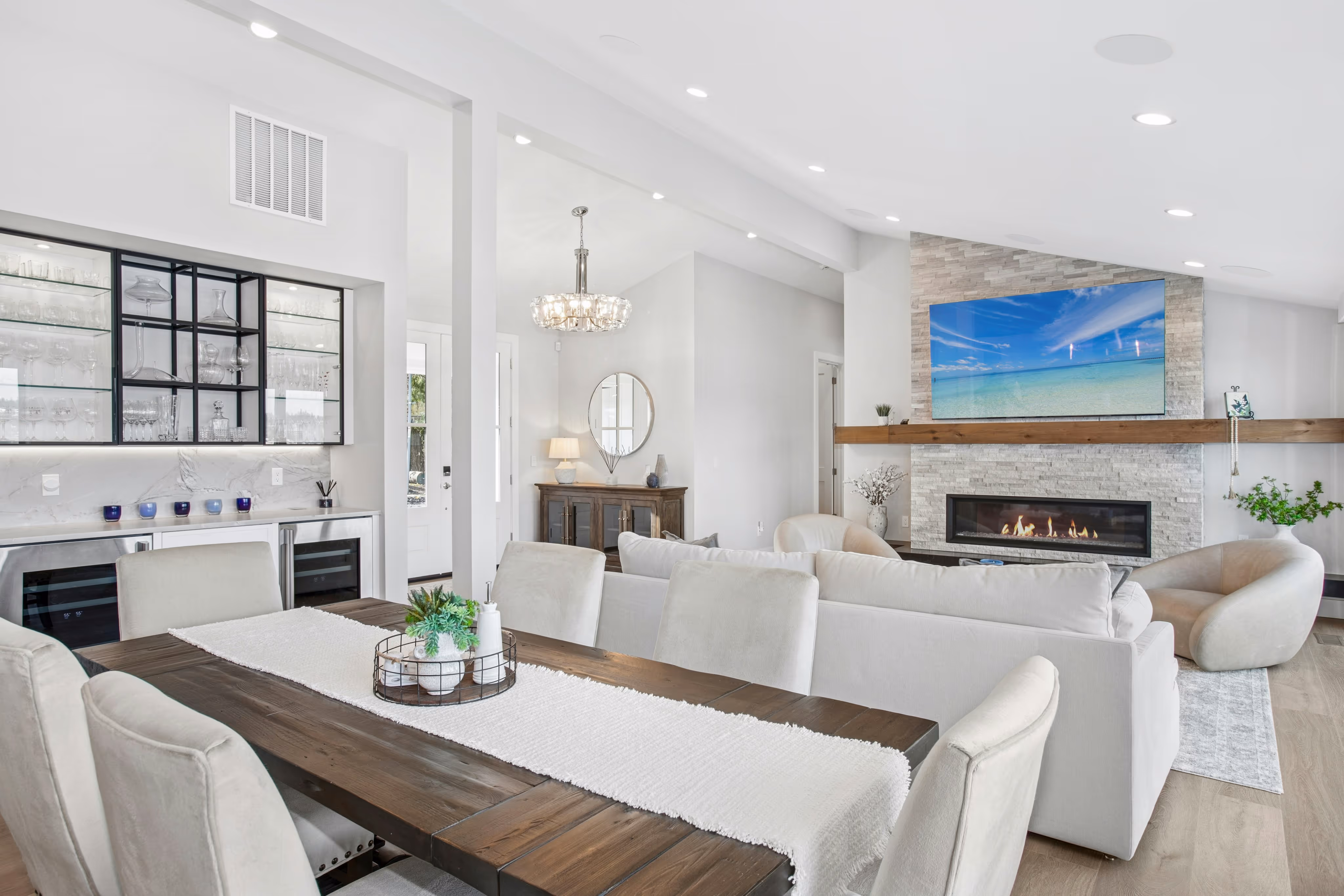 Modern open living and dining area with beige chairs, wooden dining table, white sofa, fireplace with stone surround, and wall-mounted TV displaying a beach scene.