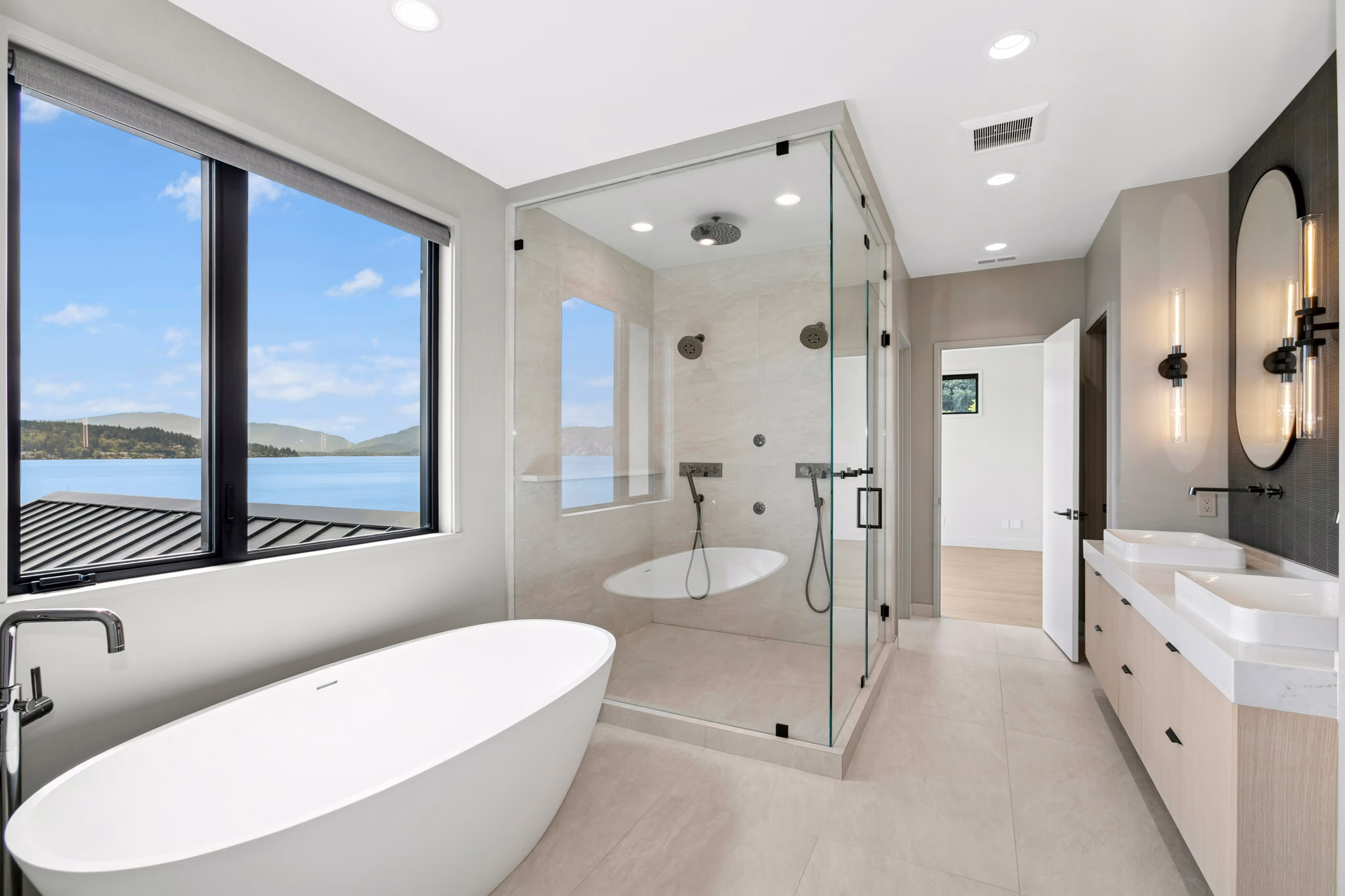 Modern bathroom with freestanding tub, glass-enclosed shower with dual showerheads, double sink vanity, and a large window overlooking a lake and mountains.