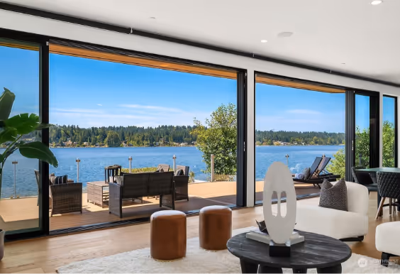 Modern living room with sliding glass doors opening to a lakeside deck featuring outdoor furniture and a scenic view of the lake and tree-lined shore.