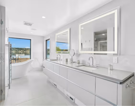 Modern white bathroom with double sinks, illuminated mirrors, freestanding bathtub, and large windows overlooking a lake.