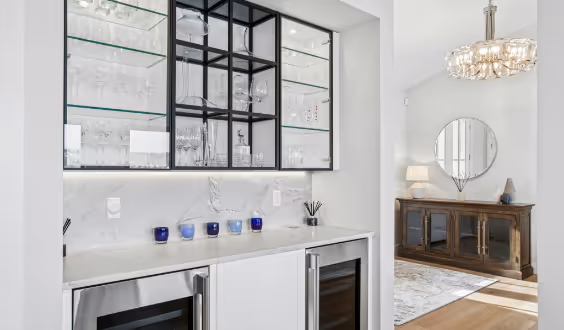 Modern home bar with glass shelves holding glassware, marble countertop with blue candles, and dual wine refrigerators below.