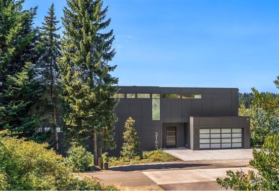 Modern two-story gray house with large windows and a triple garage surrounded by tall trees and greenery under a clear blue sky.