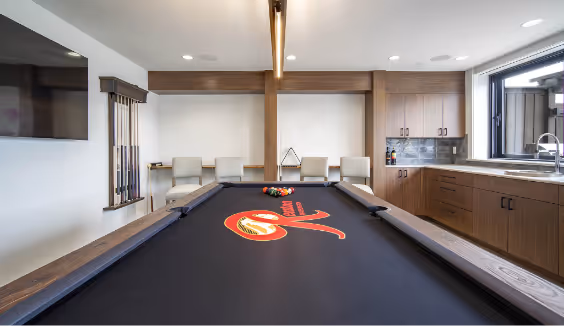 Modern game room with a black felt pool table featuring a red logo, beige chairs, mounted pool cues, a wall-mounted TV, and a kitchenette with wooden cabinets and a sink.