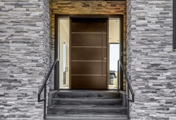 Modern dark brown front door with horizontal lines set in stone wall with gray bricks and black metal handrails on concrete steps.