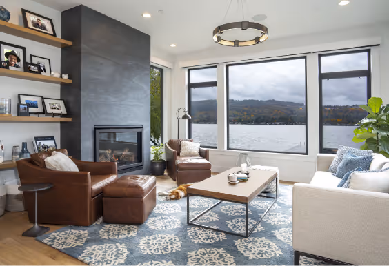 Modern living room with large windows overlooking a lake and mountains, featuring leather chairs, a white sofa, a coffee table, and a fireplace.