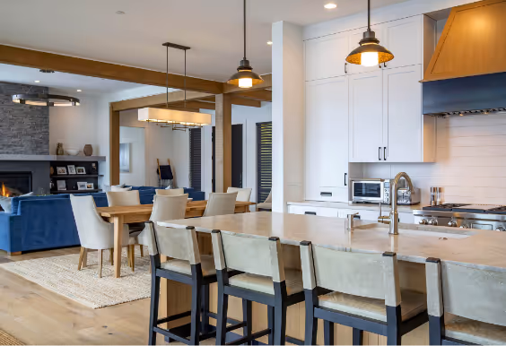 Modern open-concept kitchen with white cabinets, a large island with beige marble countertop and six upholstered bar stools, and a living area with blue sofa and fireplace in the background.