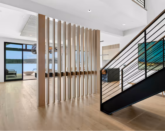 Modern living space with light wood flooring, vertical wooden slats partition, black metal stair railing, and large windows overlooking a water view.