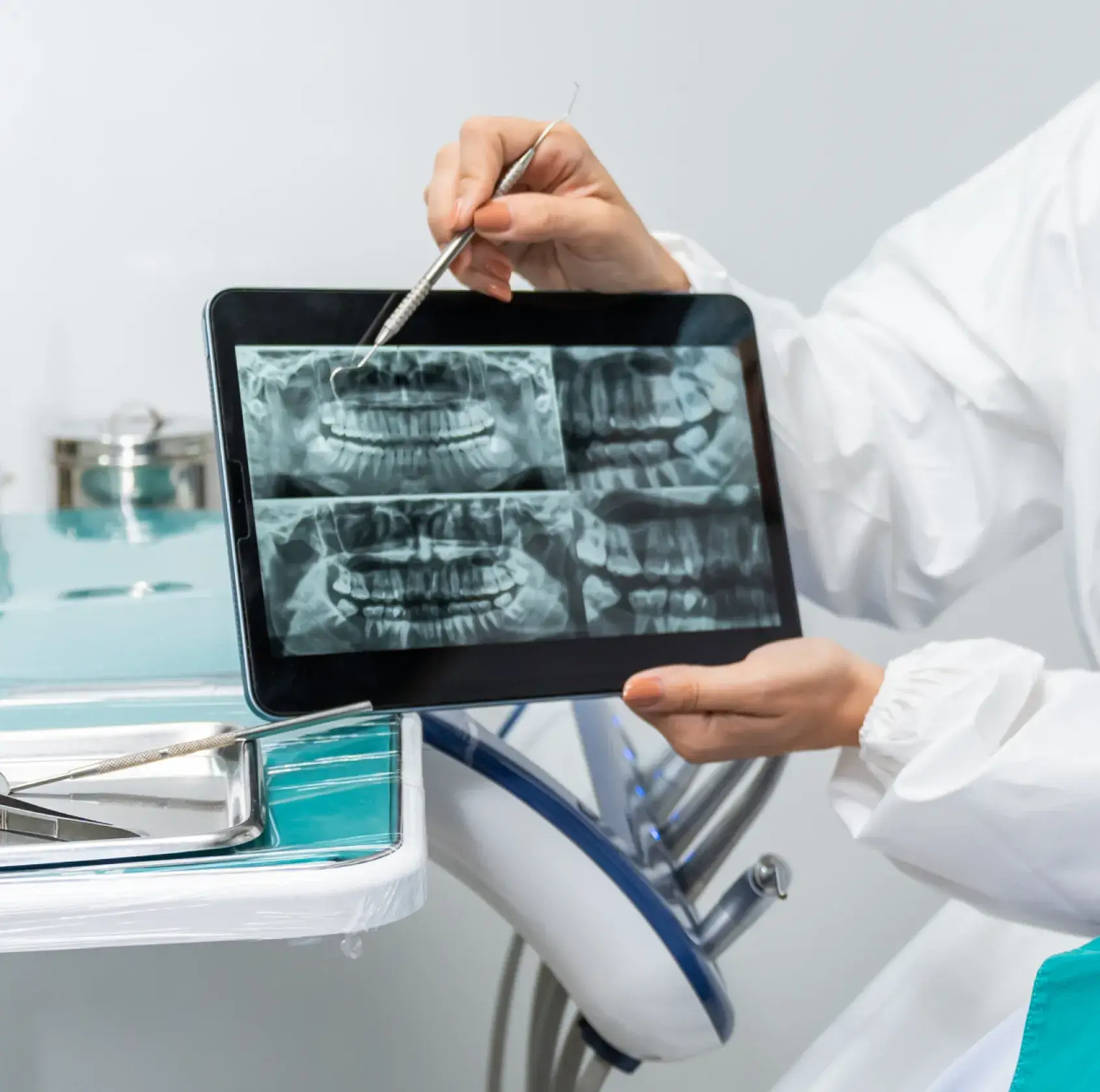 Dentist in white coat holding a tablet showing four dental X-rays and pointing at one with a dental tool.