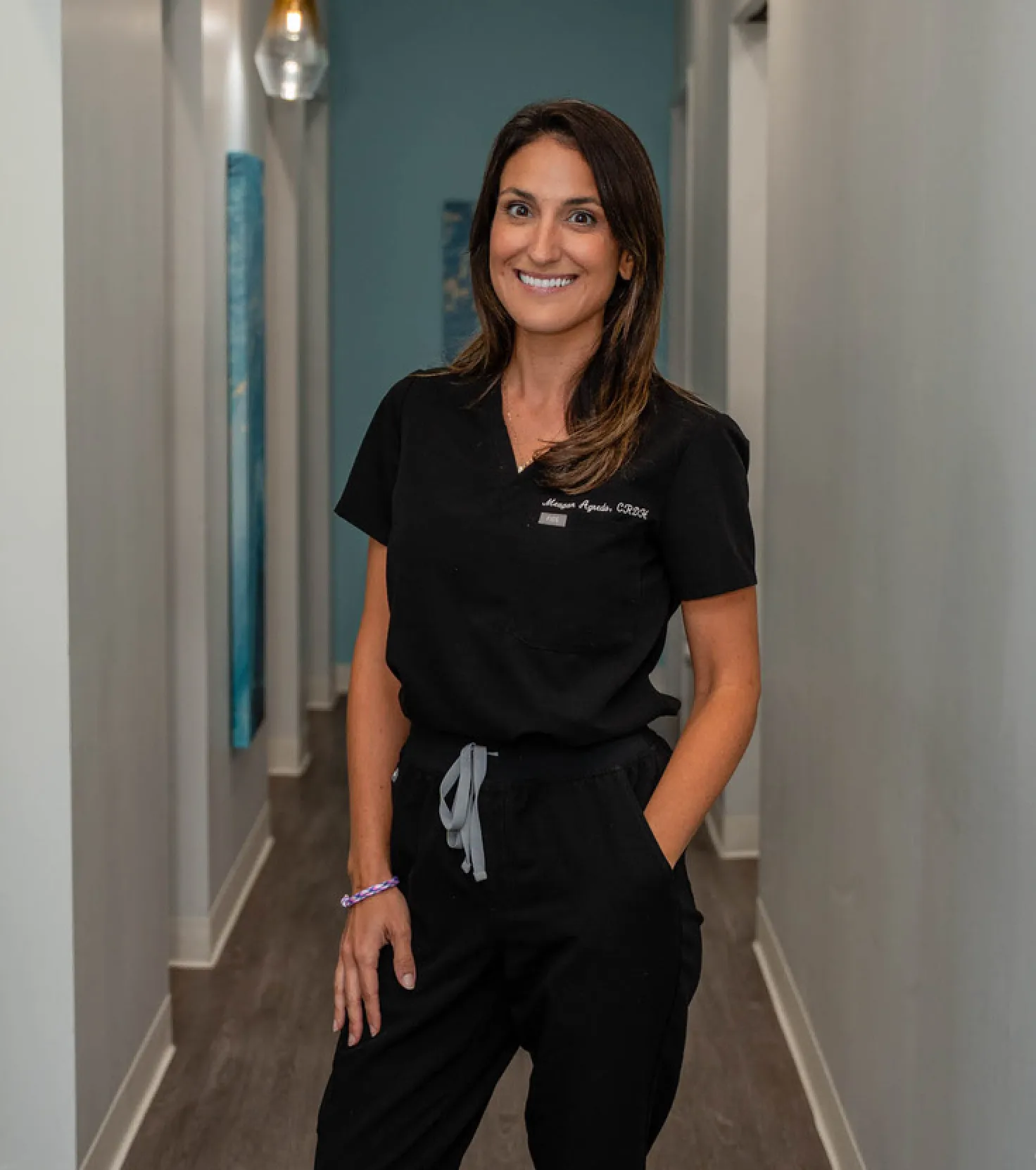 Smiling woman in black medical scrubs standing in a hallway with one hand in her pocket.