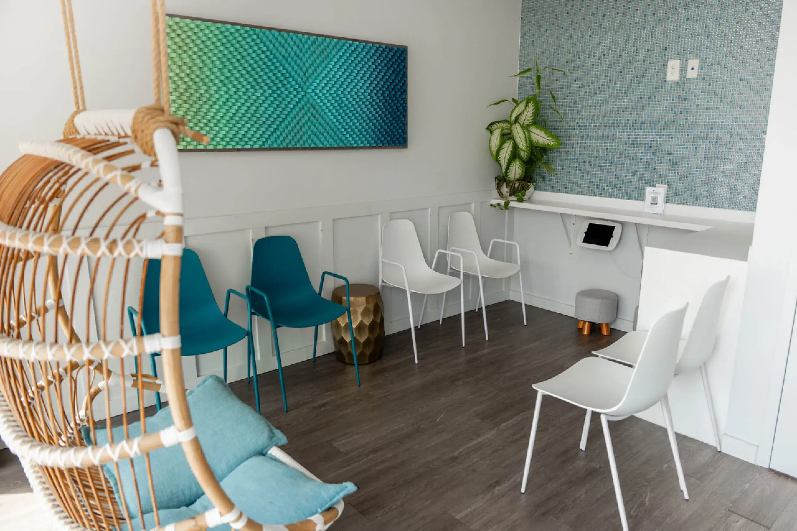 Modern waiting room with blue and white chairs, hanging wicker chair with blue cushions, green plant, and blue textured wall art.