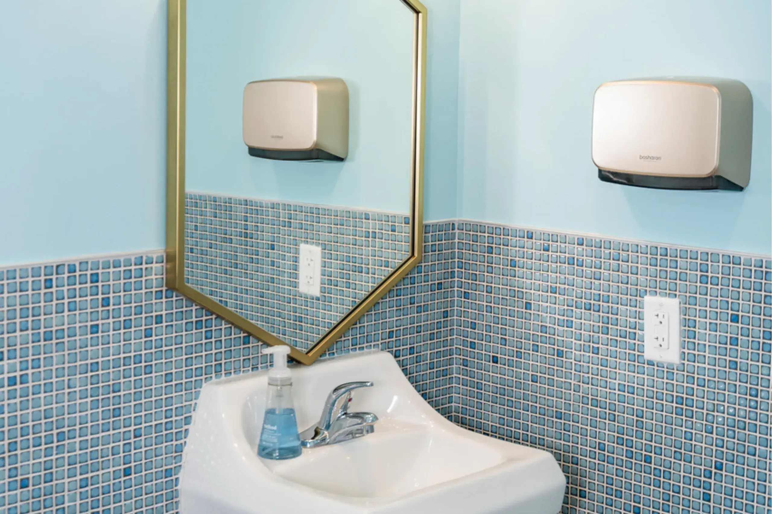 Bathroom corner with a white sink, blue tiled walls, a soap dispenser on the sink, a mounted hand dryer, and a framed mirror reflecting part of the room.