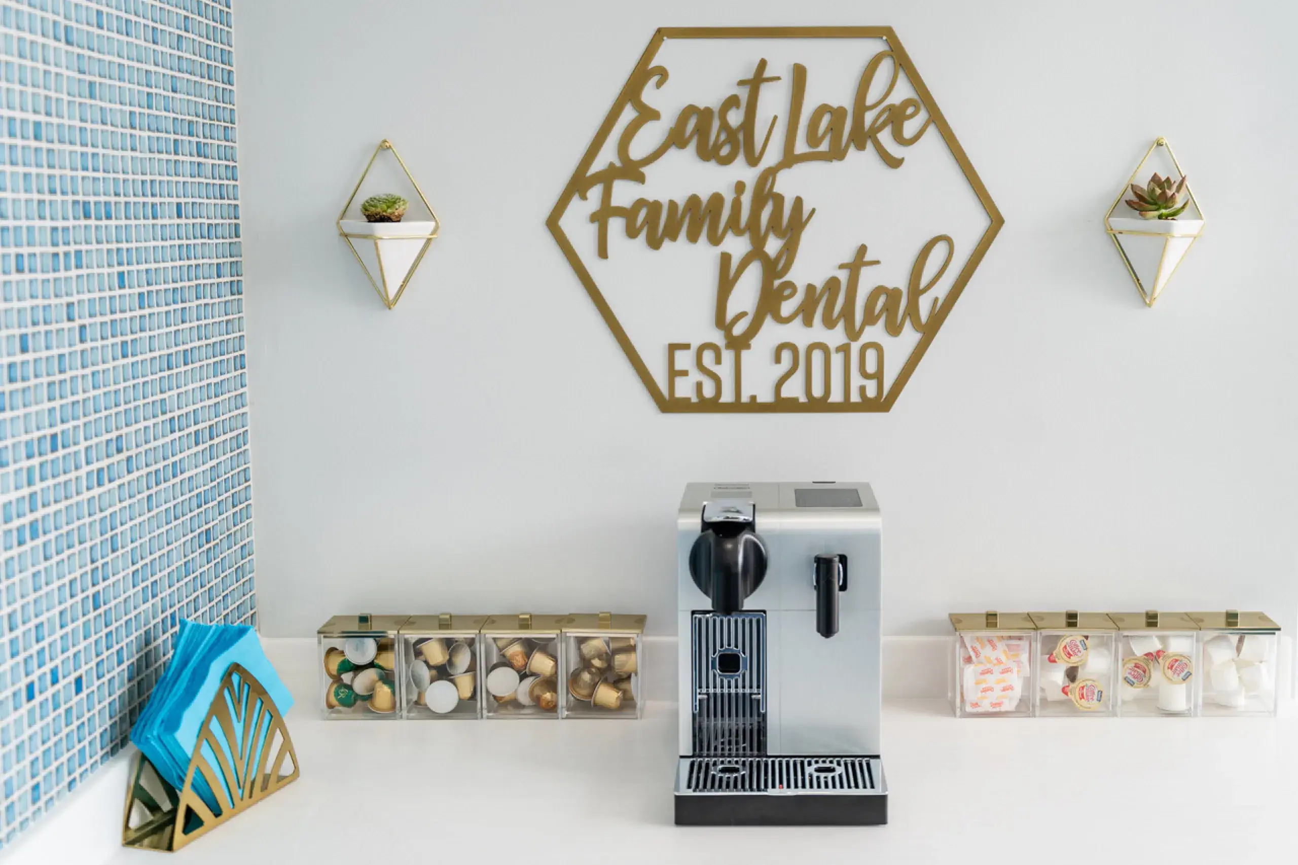 Modern coffee station with a silver espresso machine, pods, creamers, sugar packets, and blue napkins under a wall sign reading East Lake Family Dental EST. 2019.